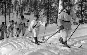 Five soldiers dressed in white hooded snow camouflage suits and traditional hats move dynamically on skis down a snowy, tree-covered slope through a Finnish forest.