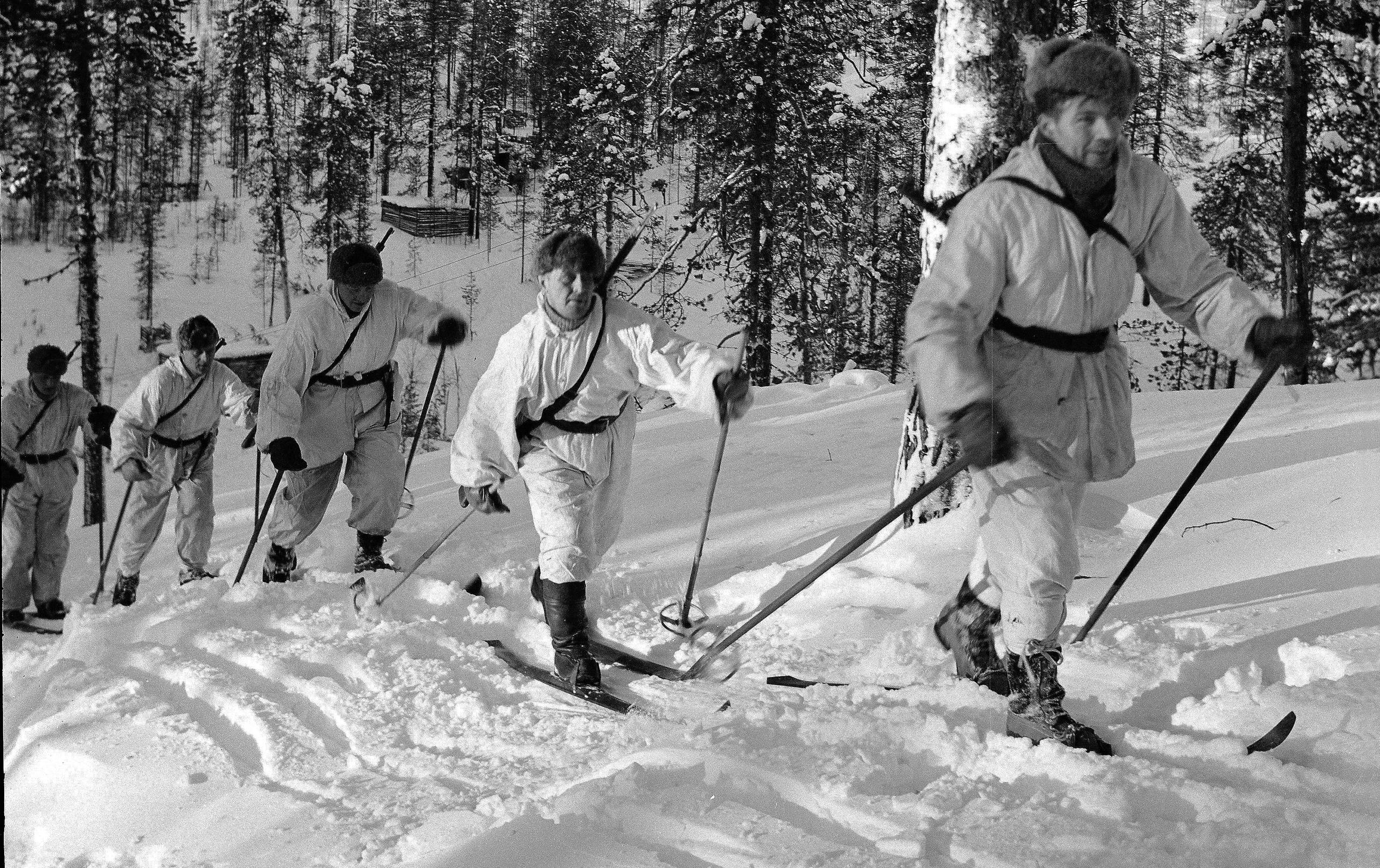 Five soldiers dressed in white hooded snow camouflage suits and traditional hats move dynamically on skis down a snowy, tree-covered slope through a Finnish forest.