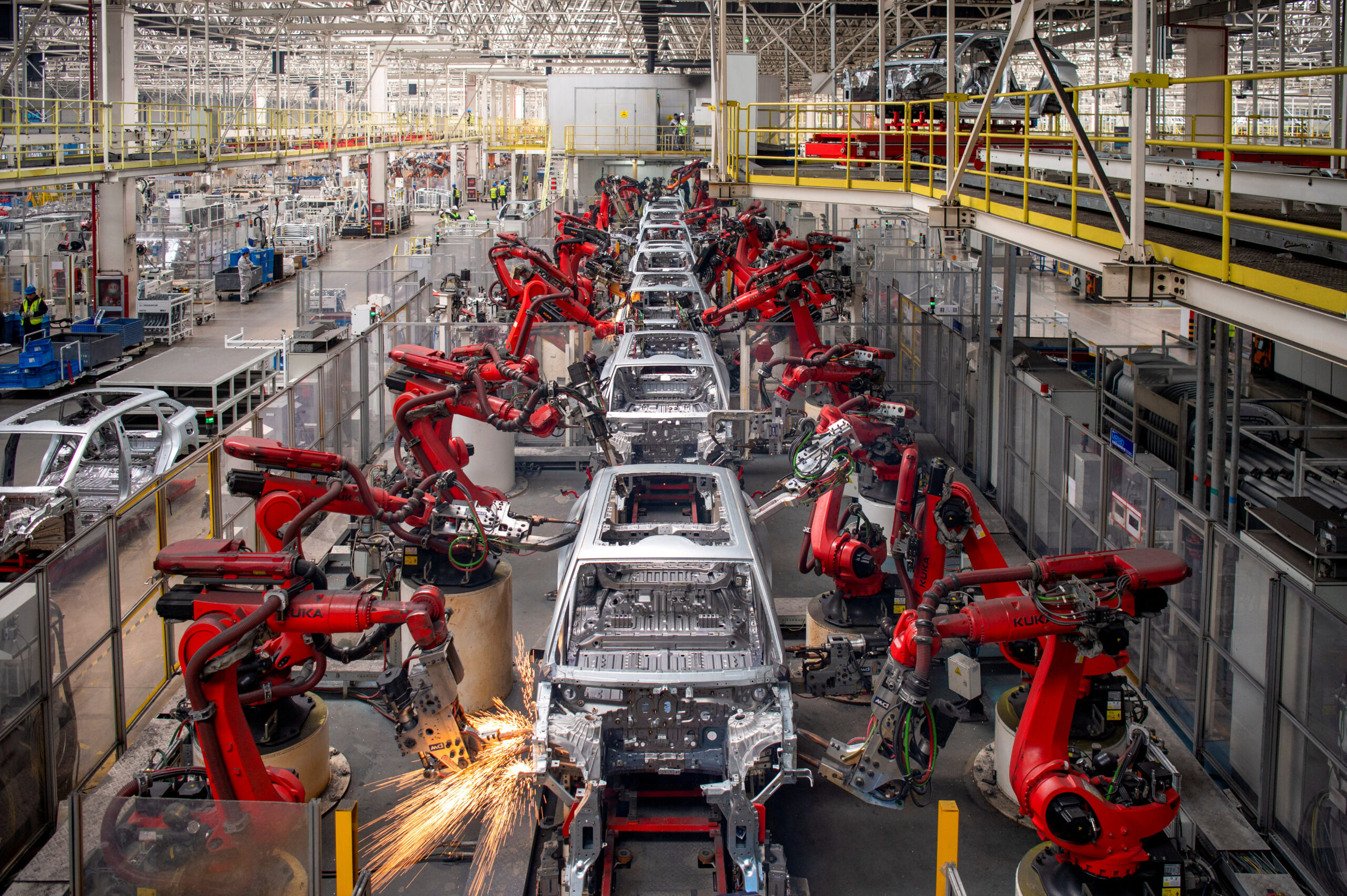 An aerial view of a modern automotive factory floor featuring a long, automated assembly line. Dozens of red KUKA robotic arms are welding car chassis in a synchronized process, with bright orange sparks flying from a point of contact in the foreground. The factory is spacious and brightly lit, with metallic car frames moving along the assembly line.