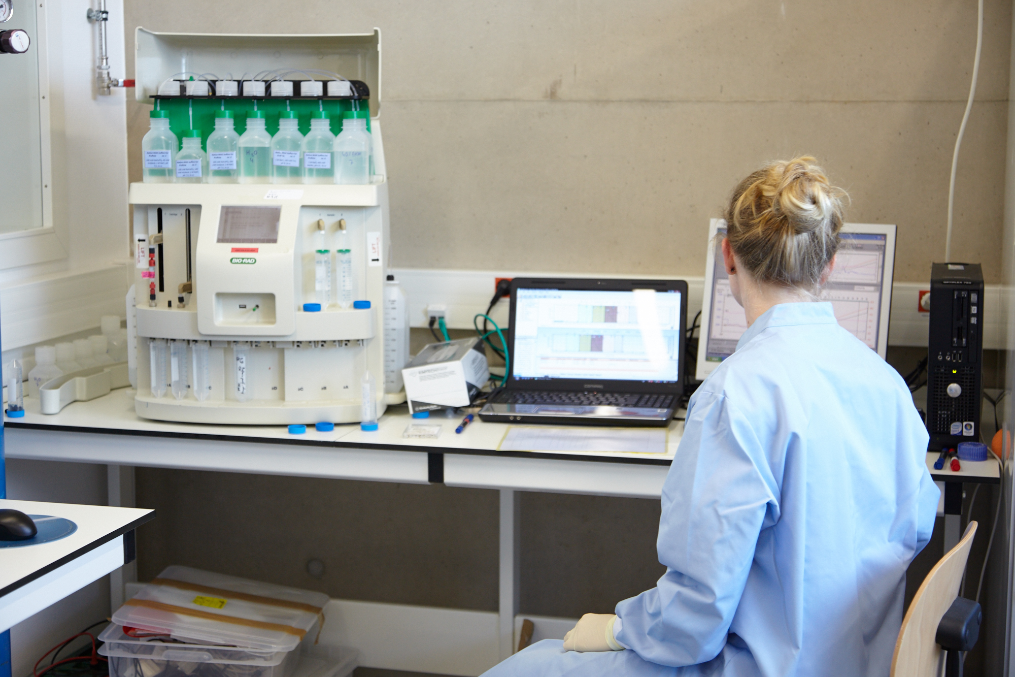 An image illustrating the unseen research effort described in the article. A female PhD researcher, often an international student according to the text, sits with her back to the camera, representing her anonymity and 'invisible serfdom' within a vast, automated academic system. She performs highly specialized data analysis, generating intellectual capital that may be claimed by local senior staff. The highly technical lab equipment signifies the resource control held by supervisors in the hierarchical system that delays defences. This visual symbolizes the academic 'labor' of international aspirants who are systematically left behind.