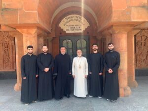 A group portrait of six Armenian Apostolic Church clergymen standing before the arched entrance of the Western Diocese Turpanjian Diocesan Building. Five men are dressed in black cassocks, while the central figure wears a white robe with a ceremonial medallion. The red sandstone architecture features intricate traditional carvings and Armenian inscriptions.