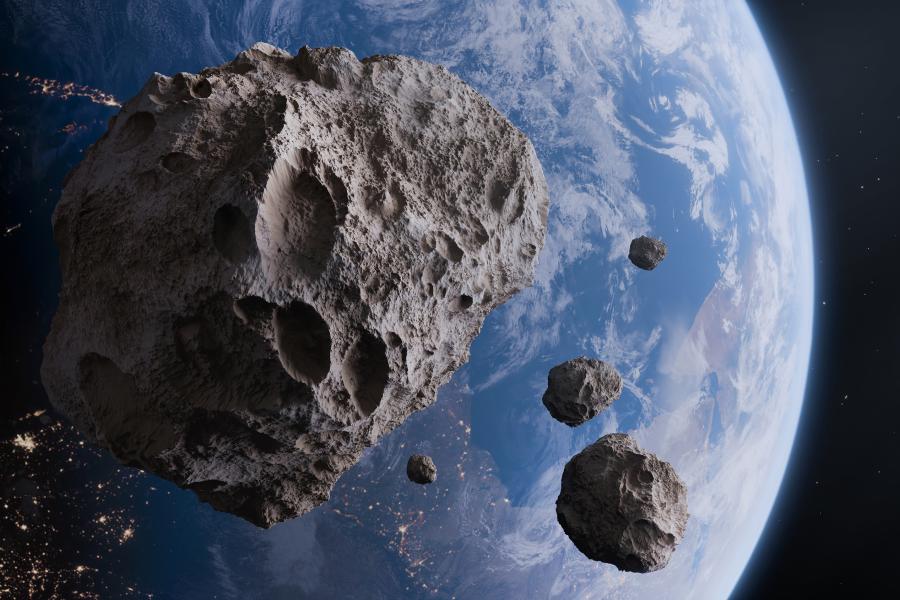 A close-up view from space showing a cluster of four asteroids orbiting Earth. The largest asteroid, on the left, is covered in significant, deep impact craters. Earth, partially lit, reveals its continents, vast blue oceans, scattered white clouds, and the distinct night-side city lights stretching across Europe and Africa along the twilight terminator. The textures of both the heavily cratered asteroid and the diverse planet are sharp and detailed.