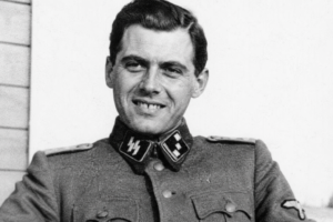A realistic black-and-white portrait of a young man in a WWII-era German SS uniform, smiling slightly while facing the camera. The image has a vintage photographic style with soft lighting, visible film grain, and a plain background emphasizing the historical military attire.