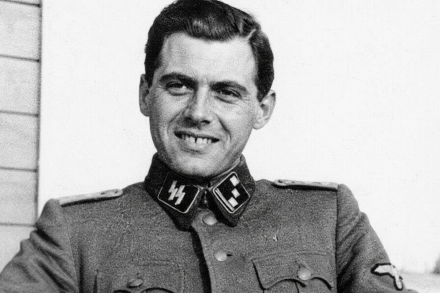 A realistic black-and-white portrait of a young man in a WWII-era German SS uniform, smiling slightly while facing the camera. The image has a vintage photographic style with soft lighting, visible film grain, and a plain background emphasizing the historical military attire.