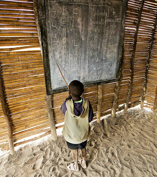 Young student standing at a chalkboard inside a rural African classroom with simple wooden walls and a sand floor, illustrating the realities of education infrastructure and access to learning in Africa