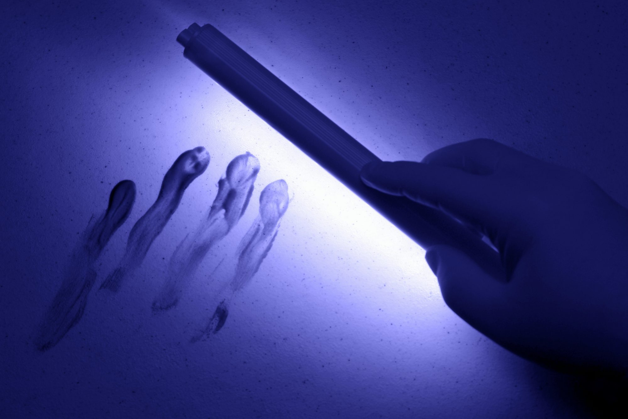 Gloved hand using a handheld UV lamp to reveal fluorescent latent handprints on a flat surface