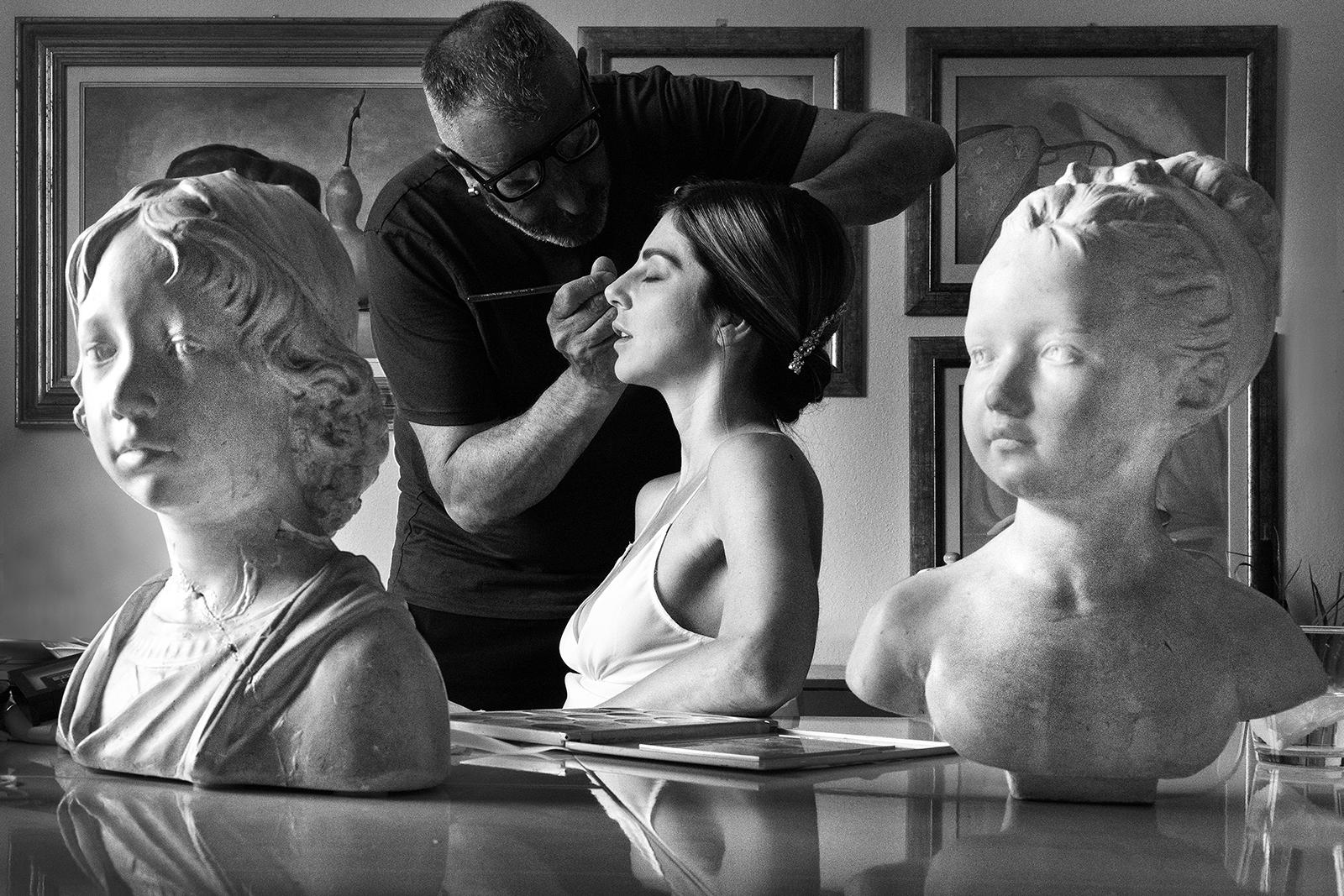 Black and white documentary photograph of a bride getting her makeup done framed by classical sculptures